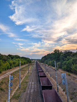 An expansive shot of a long freight train on a double-track line, with lush green trees on either side and the track disappearing into the horizon, all beneath a beautiful wide sky with dynamic clouds