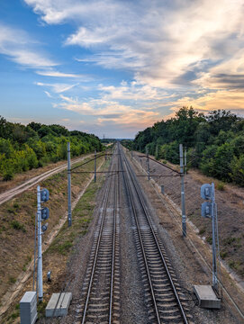 A high-angle perspective of two parallel railway tracks extending into the distant horizon, flanked by green forests and telegraph poles under a wide, partly cloudy blue sky, depicting a long journey.