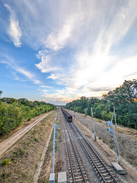 A wide-angle perspective of a cargo train on a double-track line, stretching into the horizon through a forested area, with a beautiful sky of scattered clouds and a hint of golden hour light 