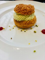 Elegant choux pastry filled with pistachio cream, artfully presented on a white plate with raspberry coulis dots and pistachio crumbles.