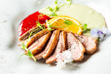 Gourmet duck breast with berry and herb sauces, orange slice, microgreens, and edible flowers on white plate. Fine dining presentation, top view