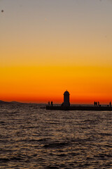 Fototapeta premium Scenic Sunset View of Lighthouse and People Silhouettes on Pier in Zadar, Croatia, Vibrant Orange and Red Sky over the Adriatic Sea Horizon, Romantic Evening Seascape