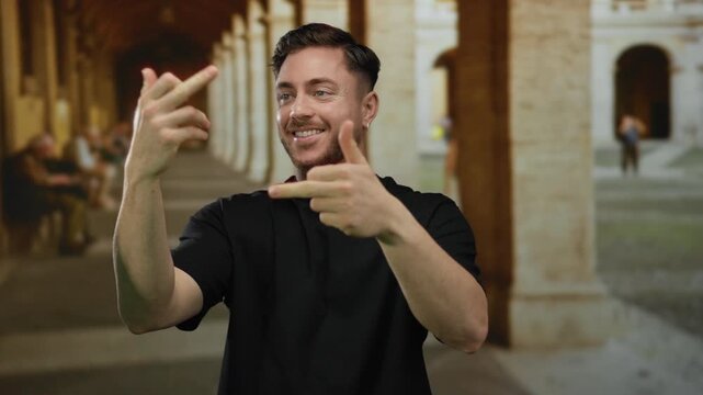 Young man with beard smiles making gestures indoors at an old university hallway with a historic backdrop, capturing a contrast between modern expression and classic architecture.