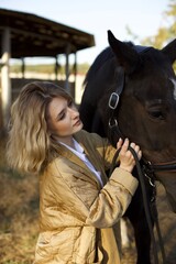Obraz premium A young woman in a cozy jacket lovingly brushes a horse's mane in a rustic barn setting. Soft light filters through, highlighting the bond between them.