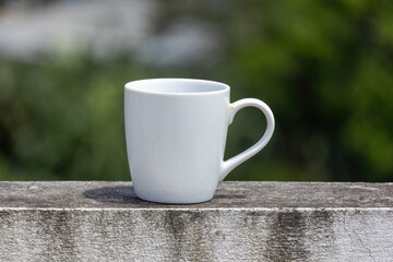 An empty white ceramic mug sits on a weathered wall with a blurred, natural background.