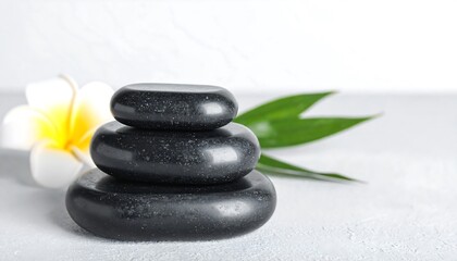 A stack of dark grey polished stones sits balanced atop a light grey surface, accompanied by a white flower and a green leaf, creating a serene and calming scene.