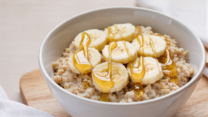 Oatmeal with banana slices and honey served in white bowl on wooden board, healthy and natural breakfast option