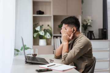 Asian man covering face with hands at desk while working from home on laptop, showing frustration and fatigue. Modern lifestyle concept of remote stress, mental health, and workplace pressure