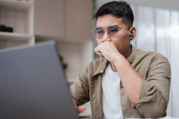 Asian man wearing glasses and earbuds looking at laptop with thoughtful expression, hand covering mouth. Remote work concept showing focus, concentration, problem solving, and professional analysis
