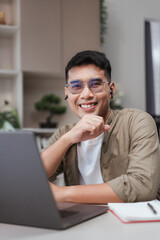 Fototapeta premium Smiling Asian man wearing glasses and earbuds looking at camera while working on laptop. Positive remote work lifestyle showing motivation, success, confidence, and professional digital workspace