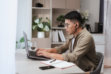 Fototapeta premium Asian man in glasses and earbuds typing on laptop at home office desk with notebook, coffee, and smartphone. Modern workspace setup for freelance projects, online work, and remote productivity