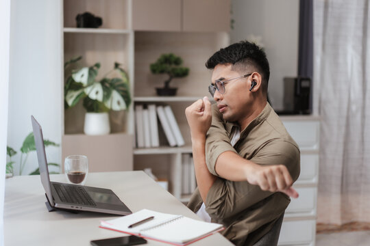Asian man stretching arms while sitting at home office desk with laptop, notebook, and coffee. Healthy remote work lifestyle concept, balancing productivity with wellness in modern workspace setup