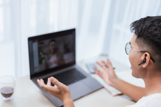 Asian man smiling with earbuds while working on laptop video call at bright modern home office desk. Great for virtual meetings, freelance productivity, and remote communication concepts