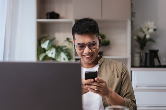 Asian man in glasses smiling while looking at smartphone, sitting at desk with laptop in bright modern home office. Great for remote work, online communication, and freelance lifestyle themes