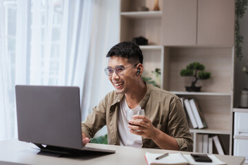 Asian man smiling while working from home, wearing glasses and wireless earbuds, holding coffee cup during video call on laptop. Perfect for remote work, online meetings, and freelance lifestyle