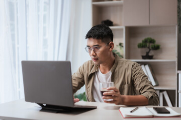 Asian man wearing glasses drinking coffee while working on laptop at modern home office desk. Relaxed telecommuting lifestyle concept, perfect for freelance projects and online work themes