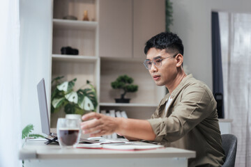 Asian man works from home at a minimalist desk, reaching for a coffee beside an open laptop and...