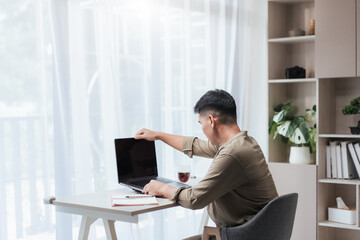 Asian man at a home desk opens his laptop to start work. A notebook, pen, and small cup sit beside him. Daylight through sheer curtains, with shelves and indoor plants in a clean, minimalist room.