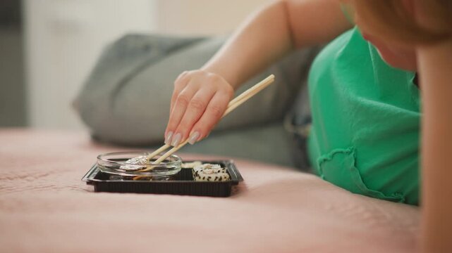 woman with polished nails lying on bed dips sushi roll into small sauce bowl using chopsticks lifting roll slowly to let excess sauce drip off calm casual setting with green shirt and pink blanket