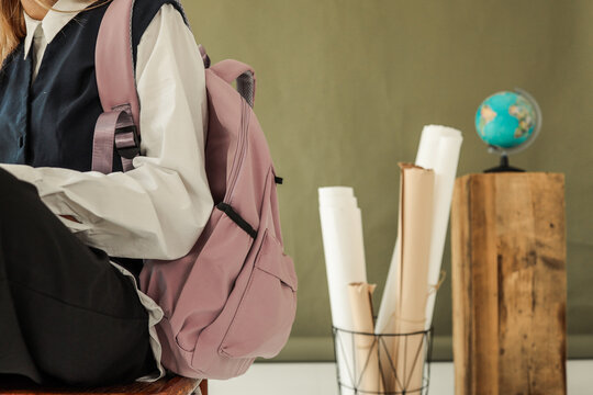 Close-up of student with pink backpack sitting on chair, school supplies with globe and rolled papers in background, education and learning concept