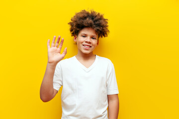 cheerful african american boy greeting and waving hand on yellow isolated background, curly...
