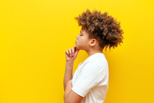 thoughtful curly african american boy standing sideways and thinking on yellow isolated background, teenager in white t-shirt looking at copy space and planning and dreaming
