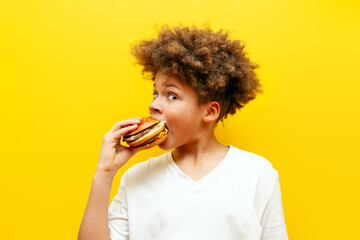 cheerful curly african american boy eating burger on yellow isolated background, hungry teenager...