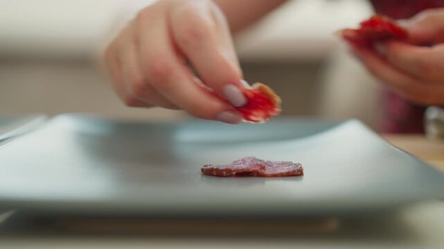 Pastry chef hands arranging single thin sausage slice on dark tray beside chopped meat pile on wooden board and whole sausage link on white table, soft natural light highlights texture and color