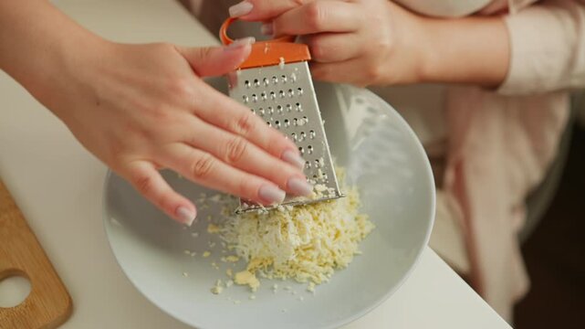 Young lady carefully grating boiled egg into plate using kitchen grater, then gently removing grater after finishing, focusing on precision and smooth cooking preparation in bright clean kitchen