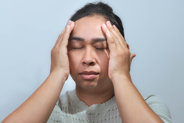 An Asian woman closing her eyes and holding her head with both hands, showing stress and pain as if...