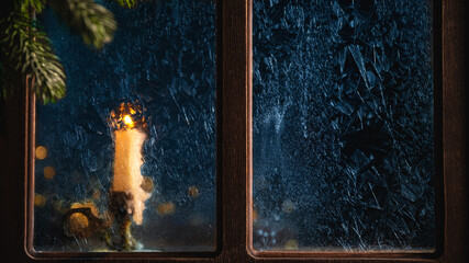 Single white candle is burning behind a frozen window during a cold winter night, creating a warm and cozy atmosphere with the fir tree branch in the foreground
