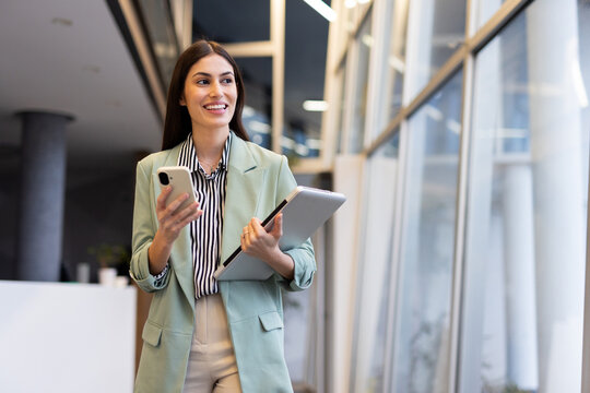 Smiling businesswoman walking in modern office hallway using smartphone and holding laptop, looking away - Powered by Adobe