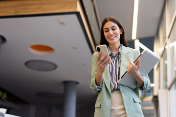 Smiling businesswoman using smartphone and holding laptop while walking in modern office hallway