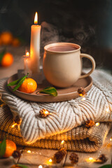 Steaming cup of hot beverage sits on a tray with a mandarin orange and lit candles, placed on a cozy knitted blanket with warm, inviting lighting, creating a perfect winter atmosphere