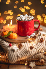 Red mug filled with hot chocolate and marshmallows resting on a tray with tangerines, surrounded by warm knit fabric and glowing christmas lights, creating a cozy winter scene