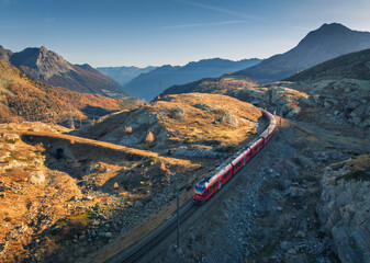 Aerial view of modern red train, alpine mountains, orange meadows, blue sky at sunset in autumn....