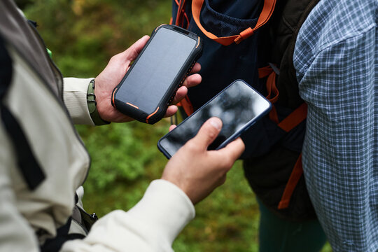 Young adult Caucasian man holding smartphone and portable solar power bank outdoors next to another , using devices while hiking or exploring forest environment