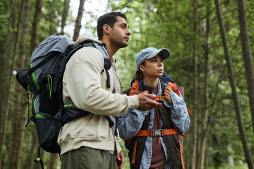 Young adult Caucasian man and young adult Asian woman hiking in forest, holding GPS device and...
