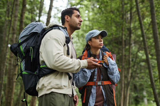 Young adult Caucasian man and young adult Asian woman hiking in forest, holding GPS device and looking ahead, wearing backpacks, surrounded by tall trees, exploring nature together