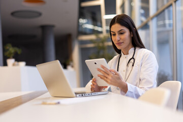 Female physician reviewing patient records on digital tablet, working in contemporary medical facility with laptop open