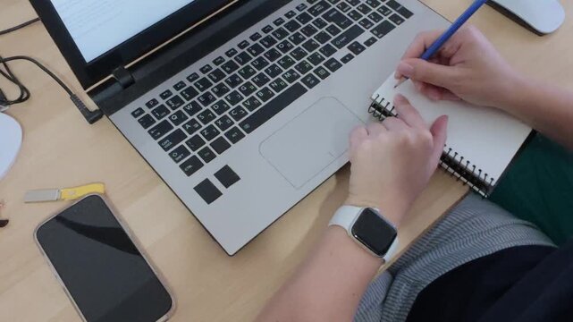 Student taking handwritten notes in a notebook during an online class or study session with a laptop
