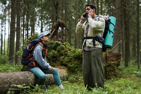 Caucasian young adult man talking on smartphone while standing in forest with hiking backpack, Caucasian young adult woman sitting on fallen tree nearby listening attentively