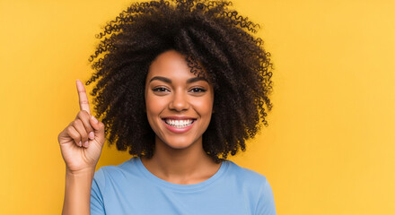 Smiling young African American woman with curly hair points her index finger upwards against a vibrant yellow background