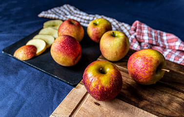 Freshly harvested apples arranged on a wooden table and a black slate tray, with vibrant red and yellow tones. Some apples are cut, revealing their white interior.