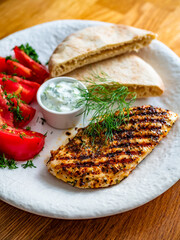 Grilled chicken breast, tomatoes, pita bread and tzatziki sauce on wooden table