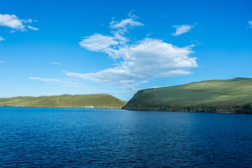 The coastal panorama of Lake Baikal under a bright blue sky, with calm water and gentle hills.