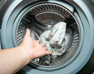 A person's hand putting a bundle of clothes into the open drum of a modern washing machine.