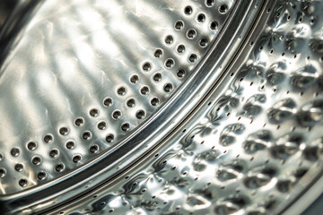A close-up of the metallic, perforated interior of a washing machine drum, showing abstract...