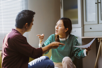 Young Asian couple arguing over paperwork at home, expressing frustration and tension as they blame each other for financial issues
