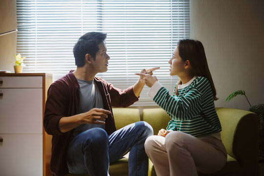 Young Asian couple arguing while sitting on a sofa at home, pointing fingers at each other, expressing frustration and tension in their relationship